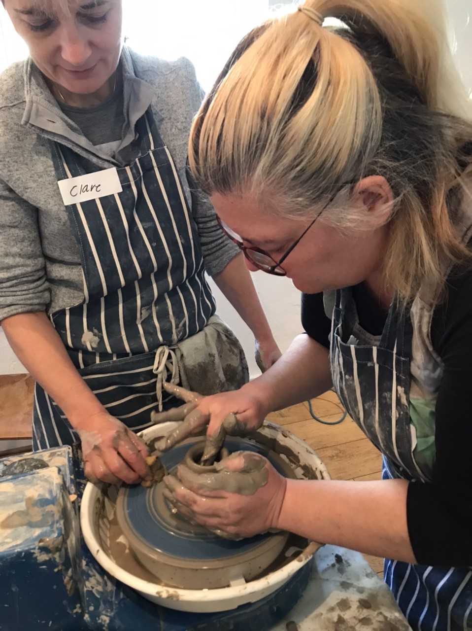 A photo from one of the taster workshops. For this workshop, the group were trying pottery. In this photo, Clare the artist is showing a member of the group how to throw a pot on a potters wheel.